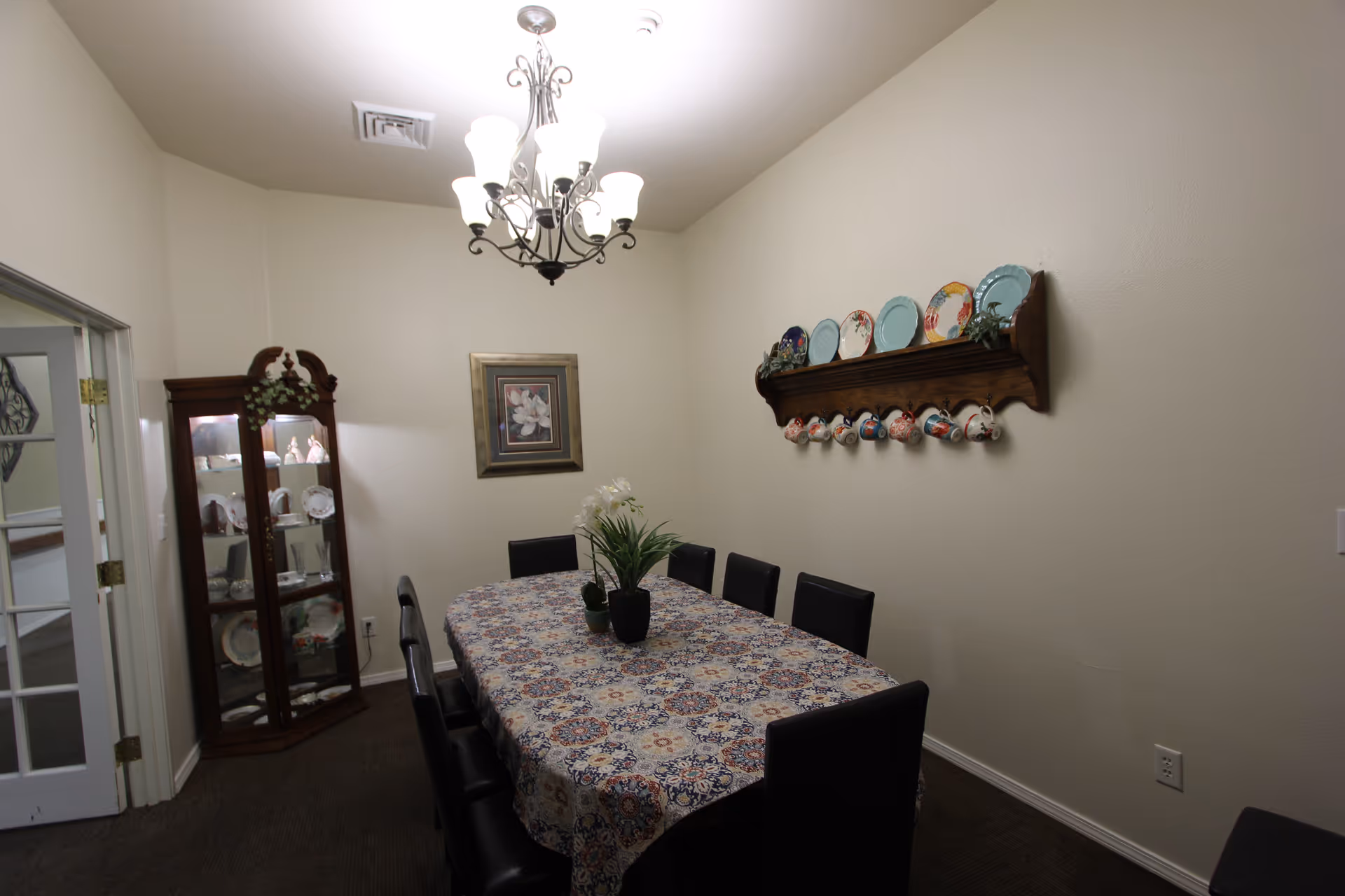 A dining room with a long table covered by a patterned tablecloth and surrounded by black chairs. On the table are two small potted plants. The room has beige walls, a chandelier with multiple lights hanging from the ceiling, a wooden display cabinet with dishes and decorative items, a framed floral picture on the wall, and a wooden shelf holding decorative plates and hanging teacups.