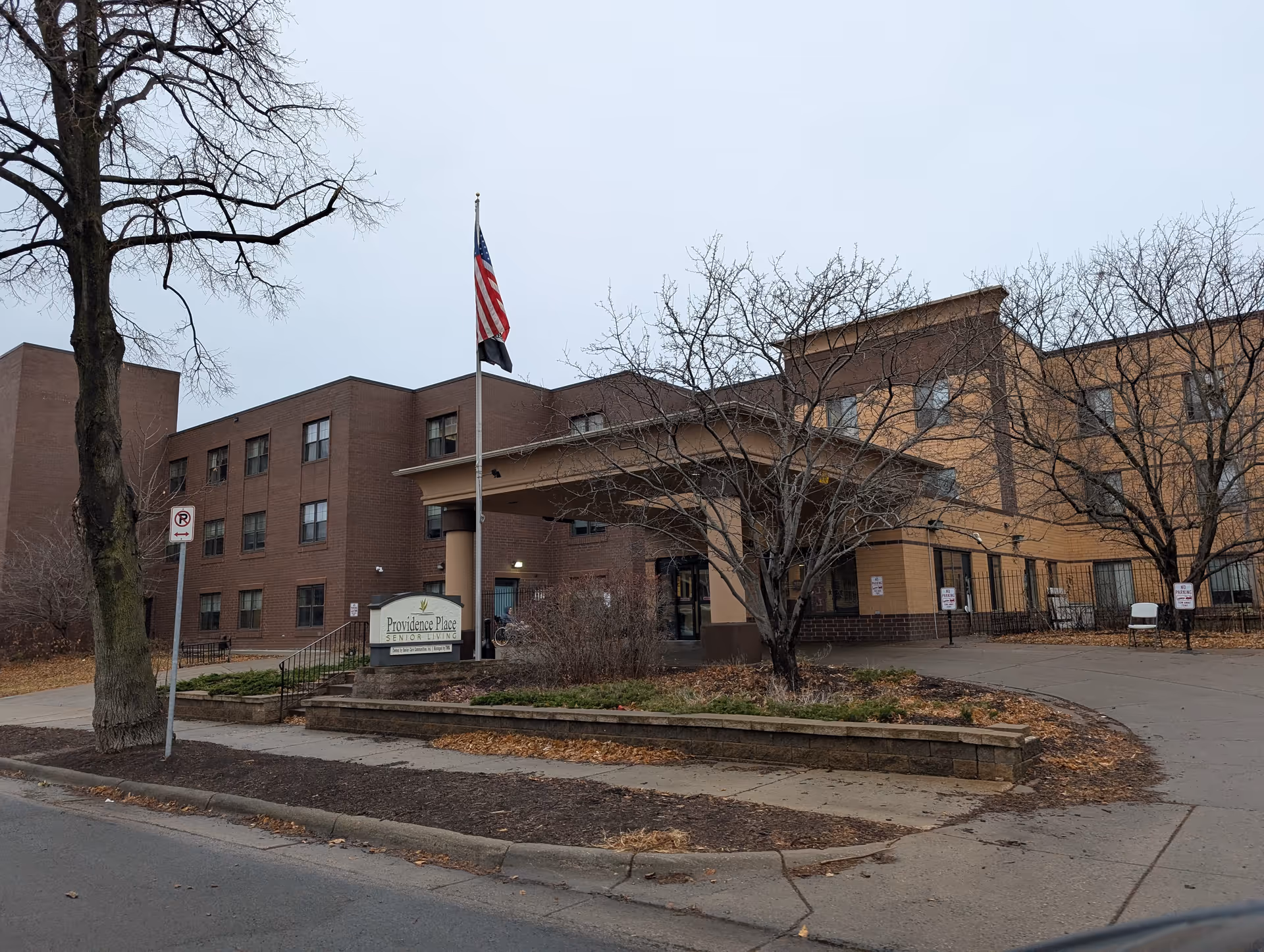 Exterior view of Providence Place Senior Living facility showing a multi-story brick building with a covered entrance, an American flag on a flagpole, leafless trees, and a sidewalk in front.