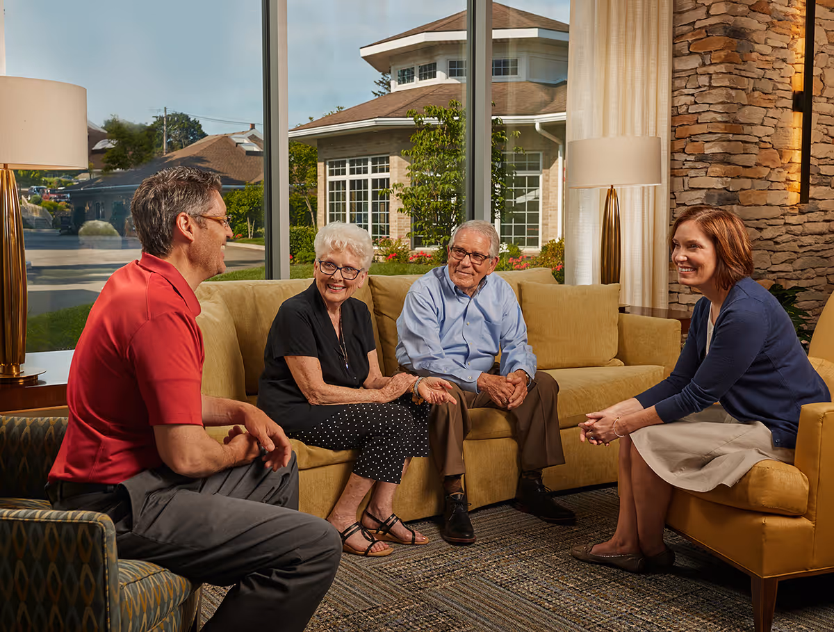 Four people sit and converse on sofas in a bright, comfortable nursing home lounge with large windows, lamps, and a stone accent wall.