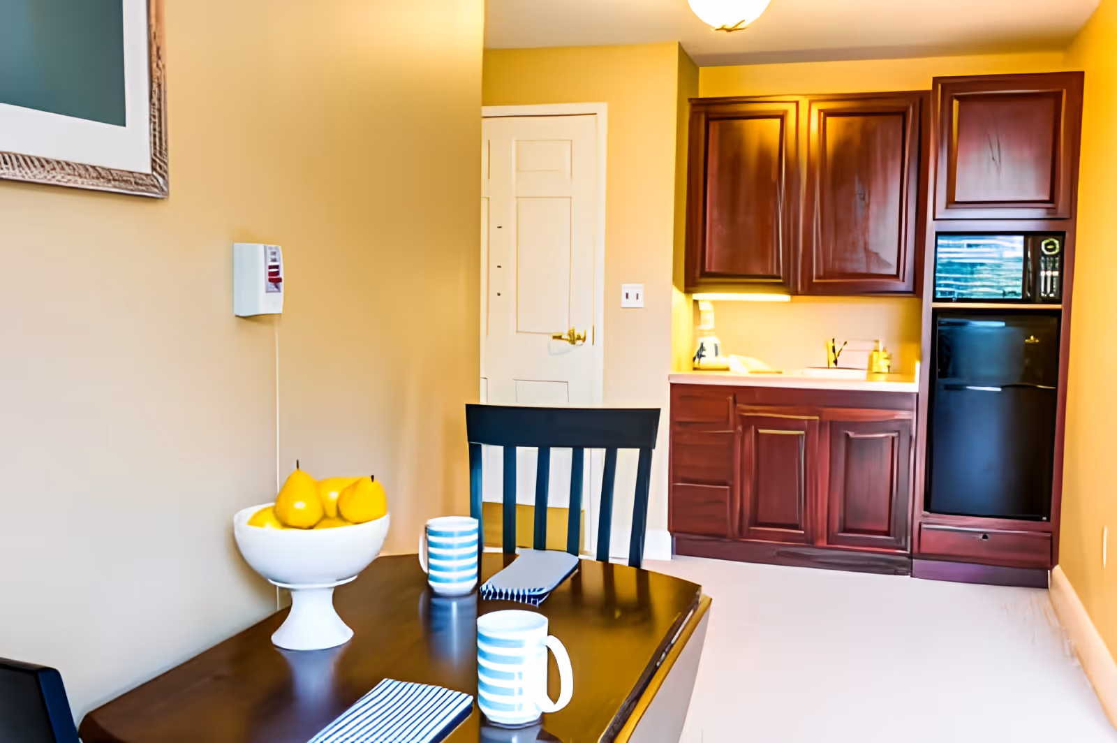 Small kitchenette with dark wood cabinets, a compact refrigerator and a dining table set with striped mugs and a bowl of yellow fruit.