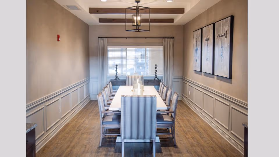 Formal dining room with a long rectangular table surrounded by upholstered chairs, wood floors, paneled walls, and artwork.