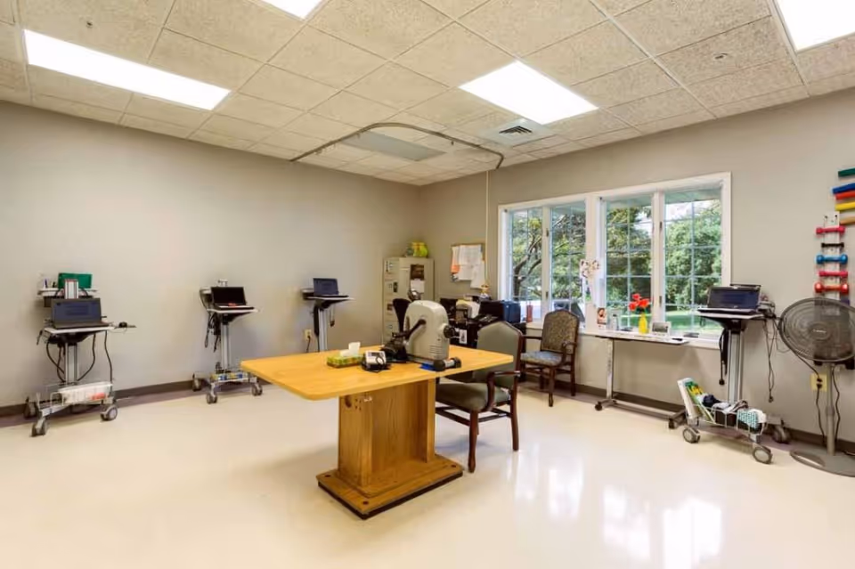 A bright room with beige walls and a white tiled ceiling featuring fluorescent lights. The room contains a wooden table with two chairs, several medical or office carts with laptops, a fan, and a window showing greenery outside. There are also some office supplies and a filing cabinet in the corner.