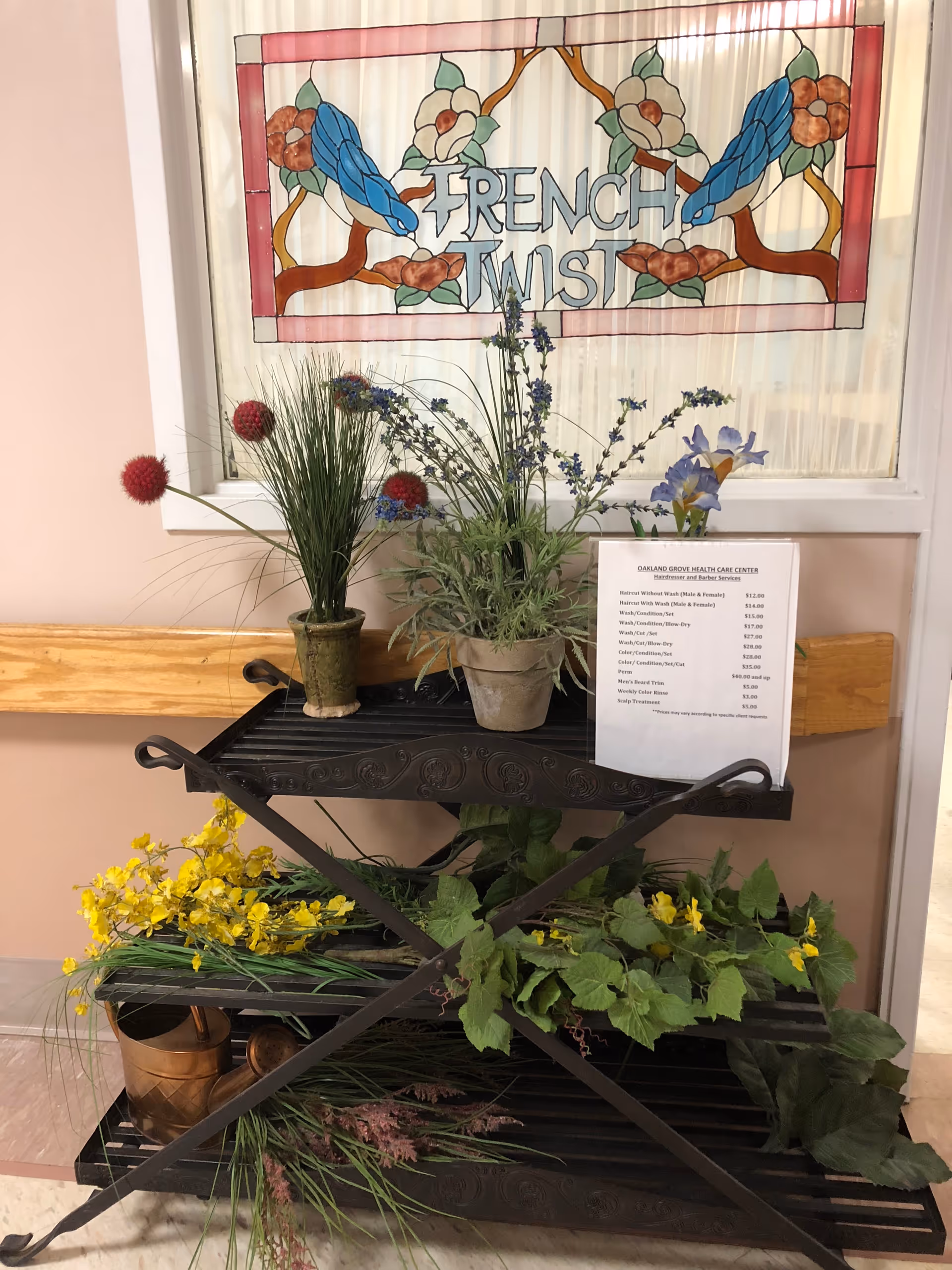 Decorative metal shelving holding potted plants and faux flowers beneath a stained-glass panel that reads "FRENCH TWIST" and a posted price list in a hallway.