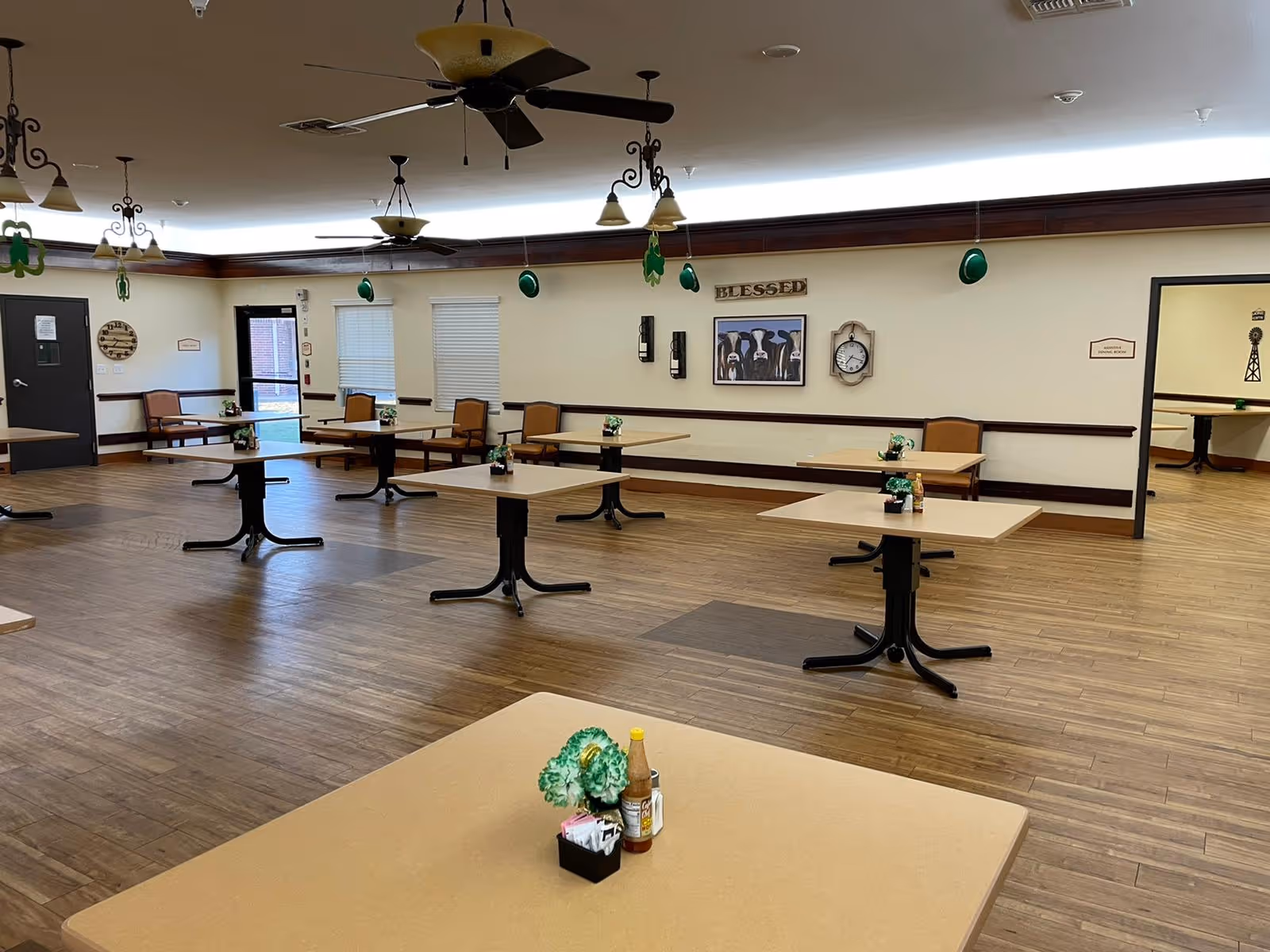 Spacious dining/activity room with multiple square tables and chairs, ceiling fans, and wall decorations including a 'Blessed' sign.