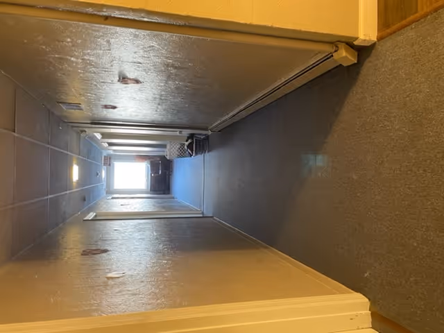 A long indoor hallway with beige walls and a gray carpeted floor. Several doors line both sides of the hallway. At the far end, there is a window letting in natural light, with a chair and some baskets visible near it.
