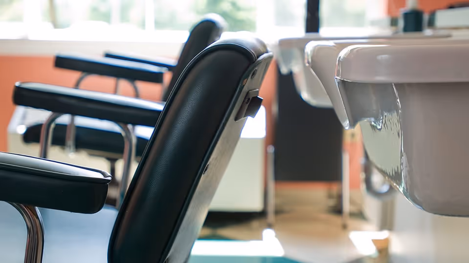 Close-up view of black padded chairs with armrests arranged in a room with large windows and a white sink visible on the right side.