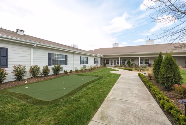 Outdoor view of TerraBella Athens facility showing a putting green on a grassy area next to a concrete walkway, with the building in the background under a partly cloudy sky.