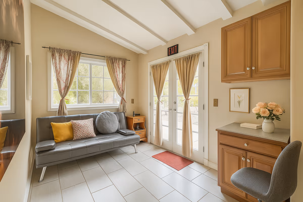 A bright and cozy interior room with a gray leather sofa adorned with three cushions in yellow, floral, and round gray designs. The room features a window with sheer floral curtains and a glass door with beige curtains tied in the middle, topped with an illuminated exit sign. To the right, there is a wooden cabinet with a vase of pink roses and a framed flower picture on the wall. A gray cushioned chair is positioned near the cabinet. The floor is tiled in light beige, and the ceiling has white beams.