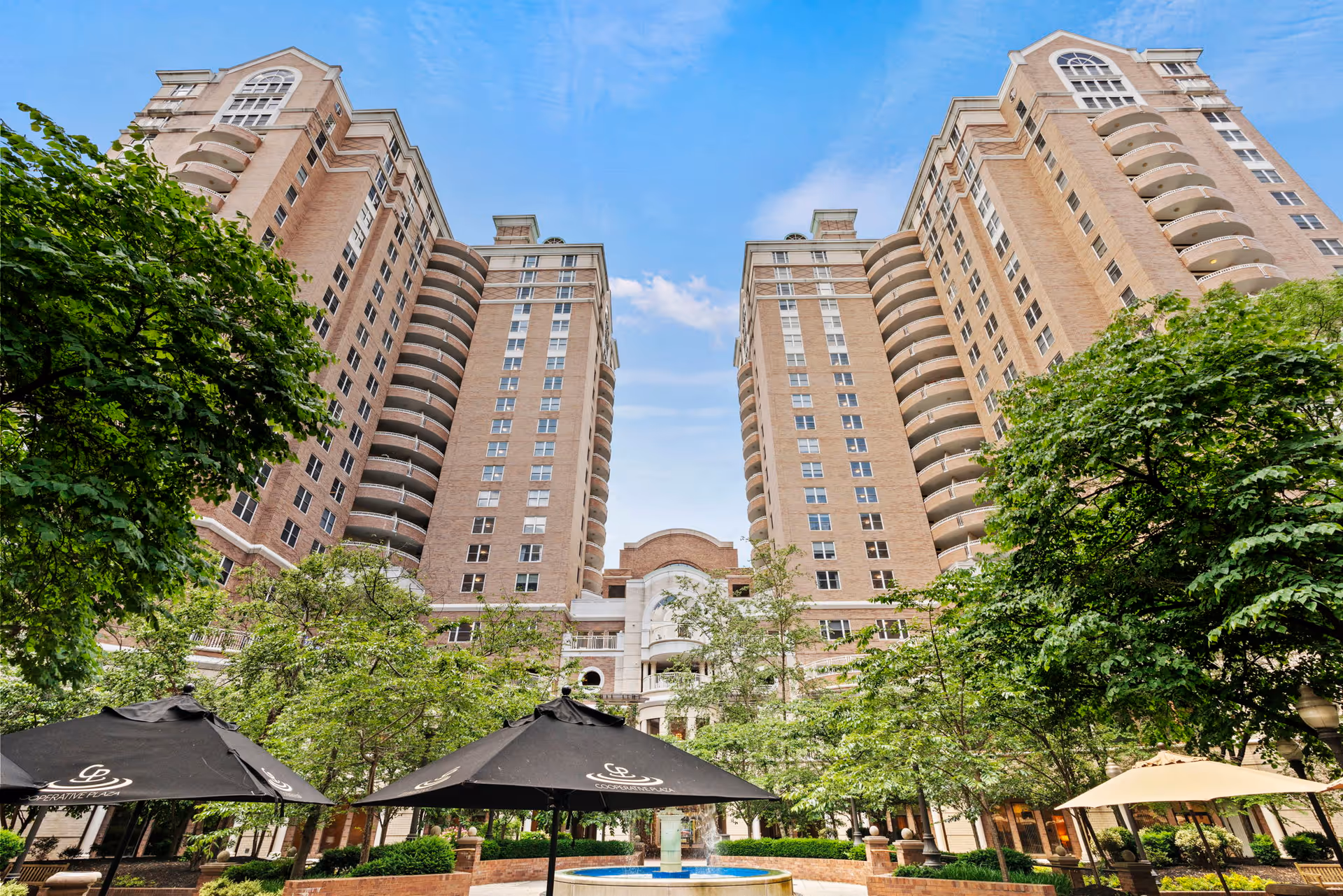 View of a tall, multi-story brick building with balconies, surrounded by green trees and outdoor umbrellas over seating areas, under a blue sky with some clouds.