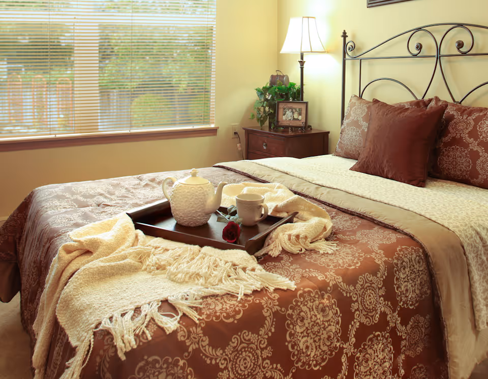 A cozy bedroom with a large bed covered in a brown and beige patterned comforter and pillows. A wooden tray with a white teapot, cup, and a red rose is placed on the bed along with a cream-colored fringed blanket. Next to the bed is a wooden nightstand with a lamp, a framed photo, and a small plant. A window with blinds lets in natural light.