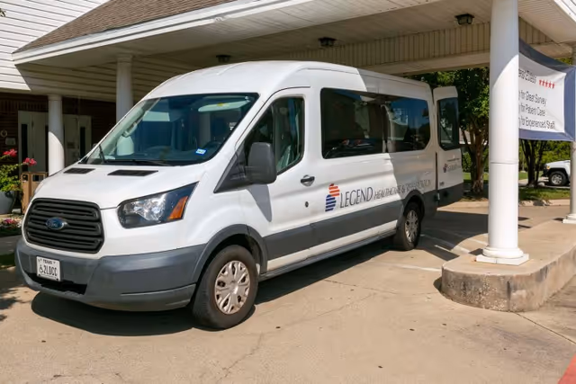 White passenger van parked under a covered driveway at a facility. The van has the logo and text 'LEGEND HEALTHCARE & REHABILITATION' on its side. The area is sunny with some greenery visible in the background.