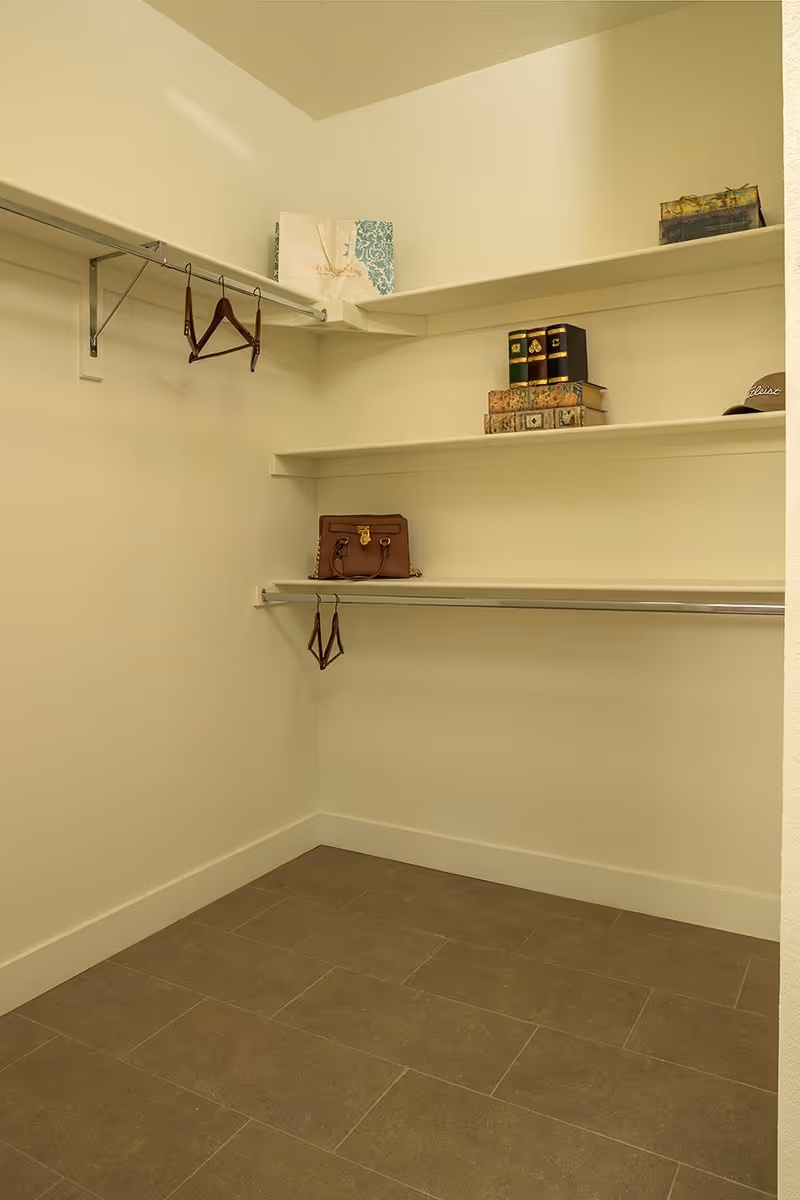 Empty walk-in closet with beige walls and brown tiled floor, featuring two hanging rods with a few wooden hangers, and three white shelves holding decorative boxes, books, a handbag, and a hat.