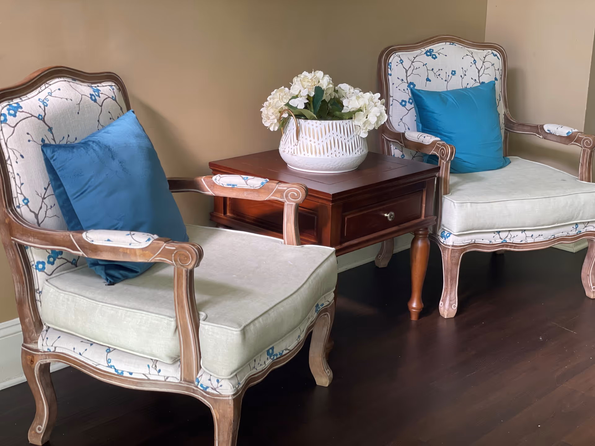 Two upholstered armchairs with wooden frames and blue floral patterns, each with a blue cushion, positioned on either side of a wooden side table with a white vase containing white flowers, set against a beige wall and dark wooden floor.