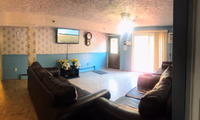 A cozy living room with two dark brown leather sofas arranged around a small black TV stand holding flower arrangements. A flat-screen TV is mounted on the wall above the stand, and a wall clock is hung nearby. The walls are decorated with floral wallpaper on the upper half and painted blue on the lower half. There is a door and a large window with vertical blinds letting in natural light. The ceiling has a textured finish with a ceiling fan and light fixture.