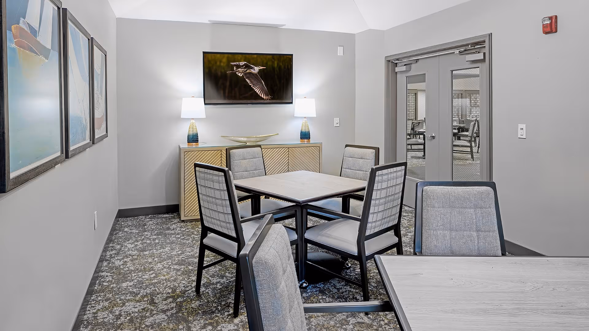 A small common area with a square table surrounded by five cushioned chairs. The room has gray walls and carpeted floor. On the far wall, there is a cabinet with two table lamps and a decorative bowl, above which hangs a framed picture of a bird in flight. To the right, double doors with glass panels lead to another room with more chairs and tables. On the left wall, three framed pictures are partially visible.