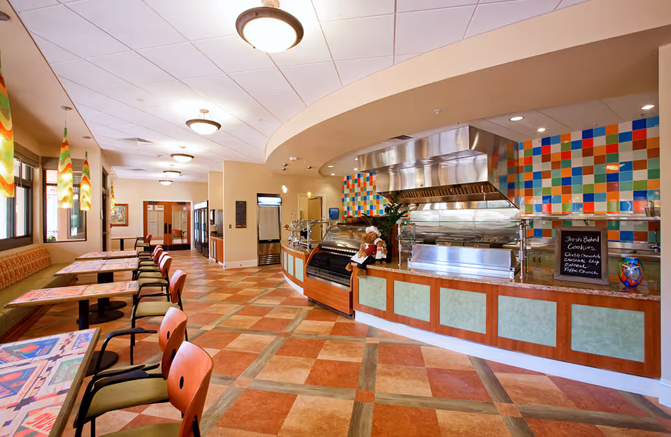 Interior view of a brightly lit dining area in a retirement community with colorful tiled walls behind a food service counter. The room features multiple tables with chairs along one side, pendant lights hanging from the ceiling, and a tiled floor with a geometric pattern. A small chalkboard sign on the counter advertises fresh baked cookies.