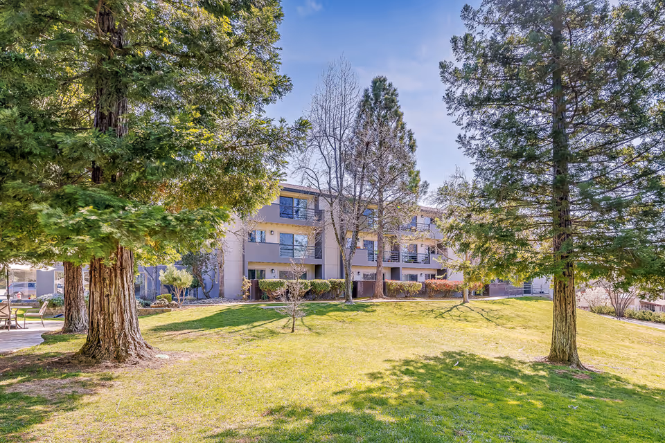 Grassy lawn and large trees in the foreground with a three-story retirement center building in the background.
