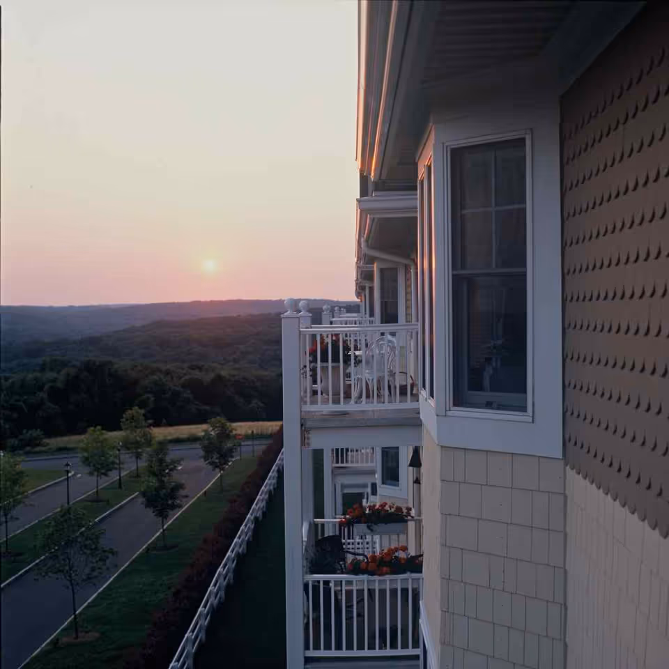 View of a senior living facility's balconies at sunset with a scenic landscape of trees and hills in the background. The balconies have white railings and some have flower pots and chairs.