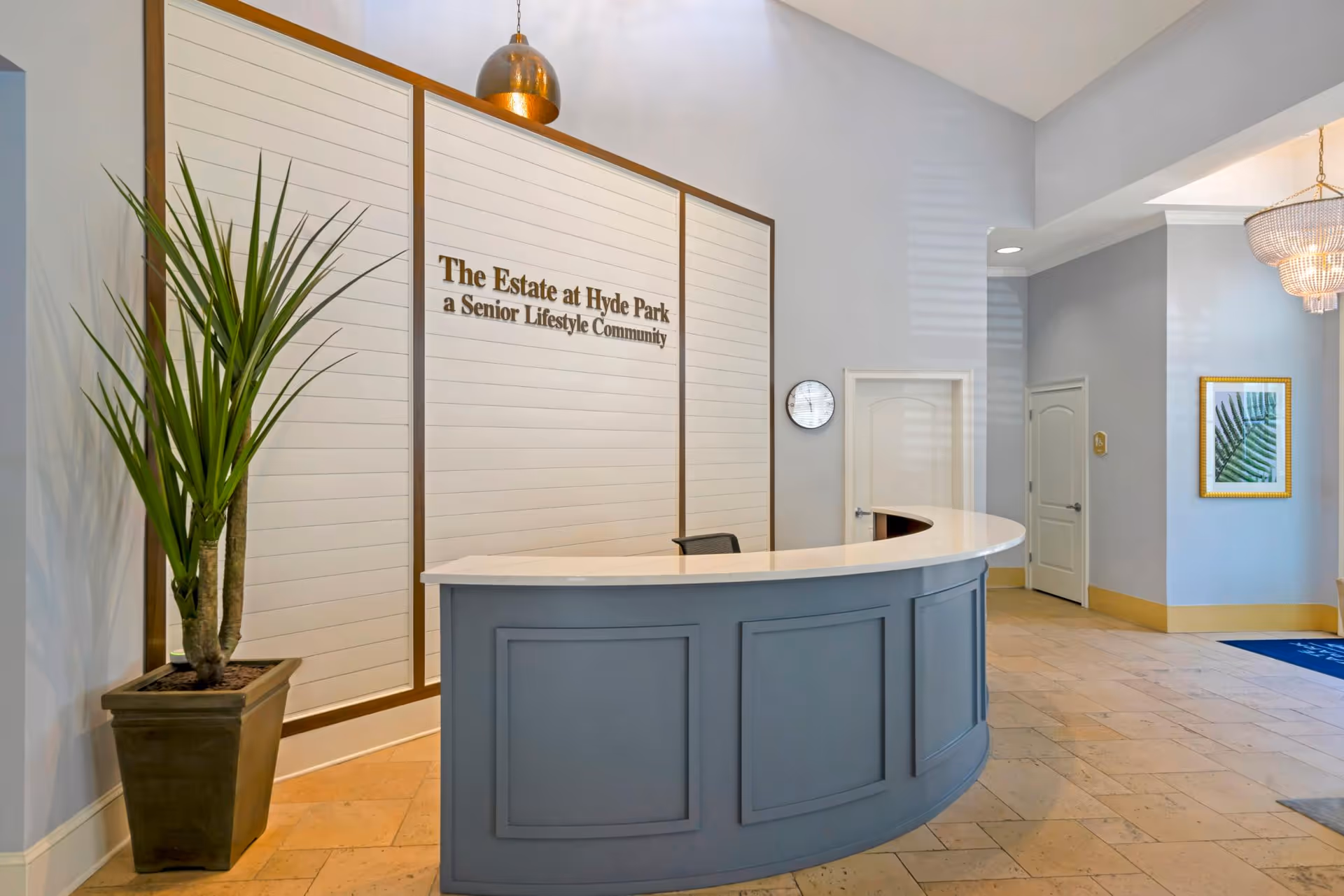 Reception area with a curved blue desk and a white countertop. Behind the desk is a wall with the text 'The Estate at Hyde Park a Senior Lifestyle Community'. There is a tall potted plant to the left of the desk, a clock on the wall, two closed doors, a framed picture of a green leaf, and a chandelier hanging from the ceiling.