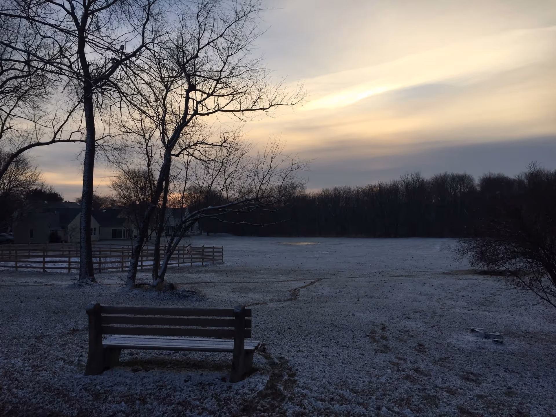 A snowy outdoor scene at dusk with a wooden bench in the foreground, leafless trees, a fenced area, and houses in the background under a cloudy sky.