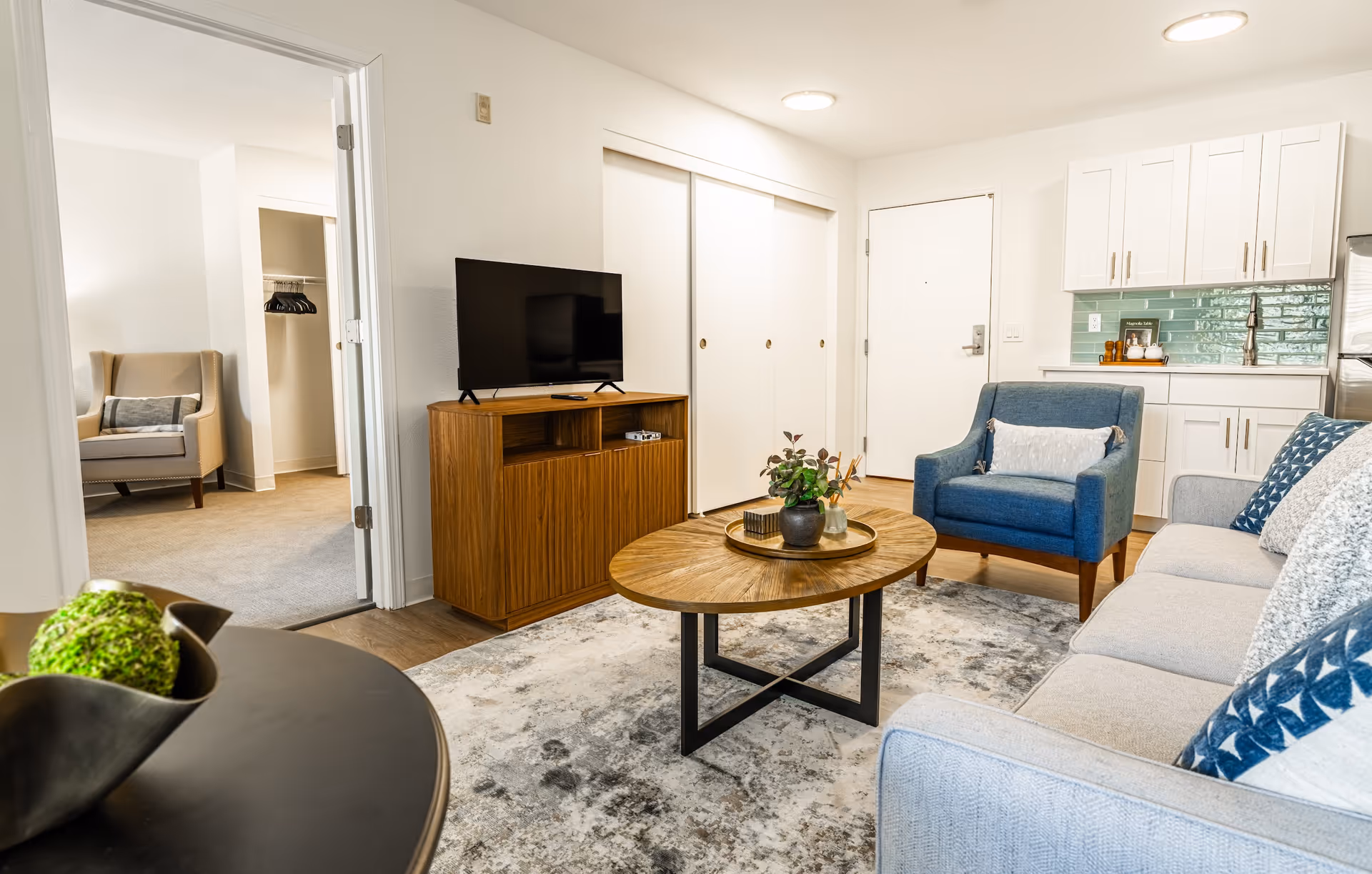 A bright and modern living room in a senior living facility featuring a light gray sofa with blue and white pillows, a blue armchair, a round wooden coffee table with decorative items, a wooden TV stand with a flat-screen TV, and an open doorway leading to a bedroom with a beige armchair and closet. The kitchen area with white cabinets and a green tiled backsplash is visible in the background.