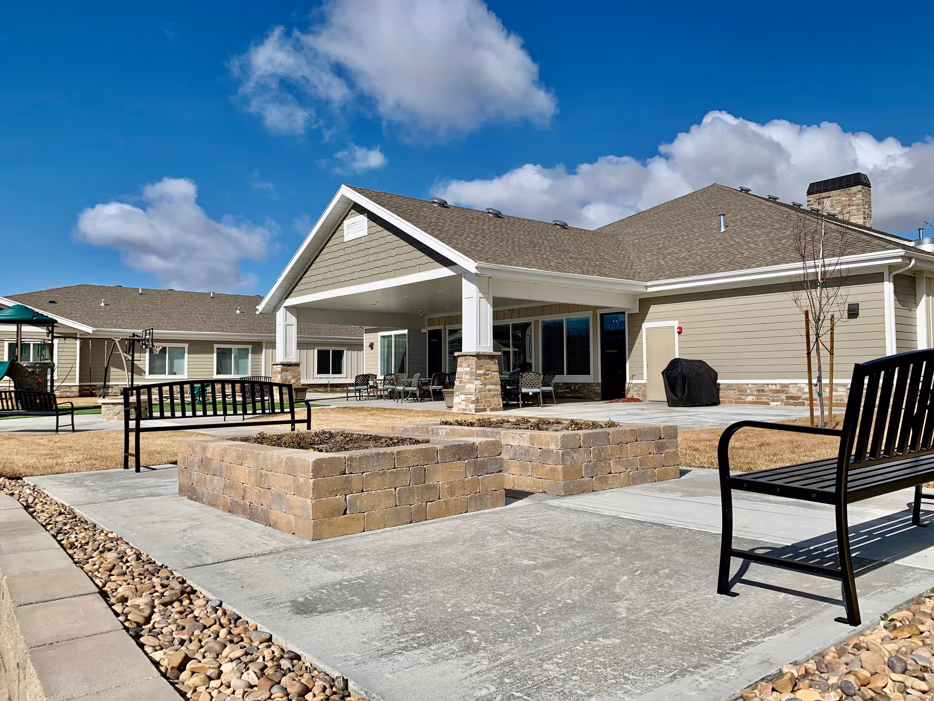 Outdoor courtyard of a senior living facility with benches, raised brick planters, and a covered patio attached to the building under a blue sky.