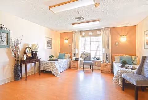 Bright memory-care bedroom with two twin beds, bedside tables, chairs, and a large window with curtains.