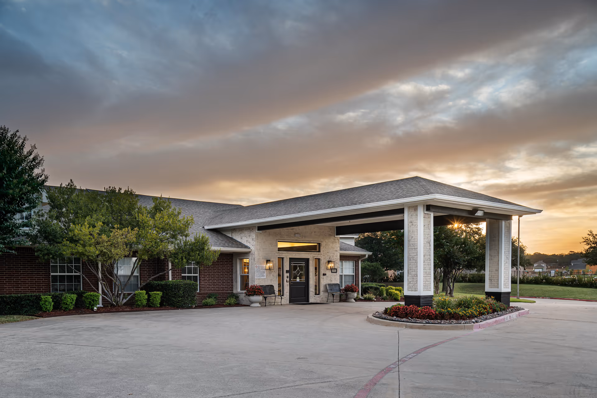 Exterior view of a single-story senior living facility building with a covered entrance, surrounded by landscaped greenery and a cloudy sky at sunset.