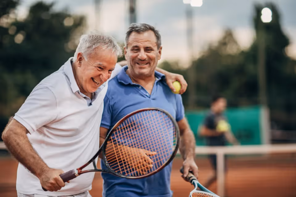 Two older men play tennis on an outdoor court, smiling as they hold rackets.