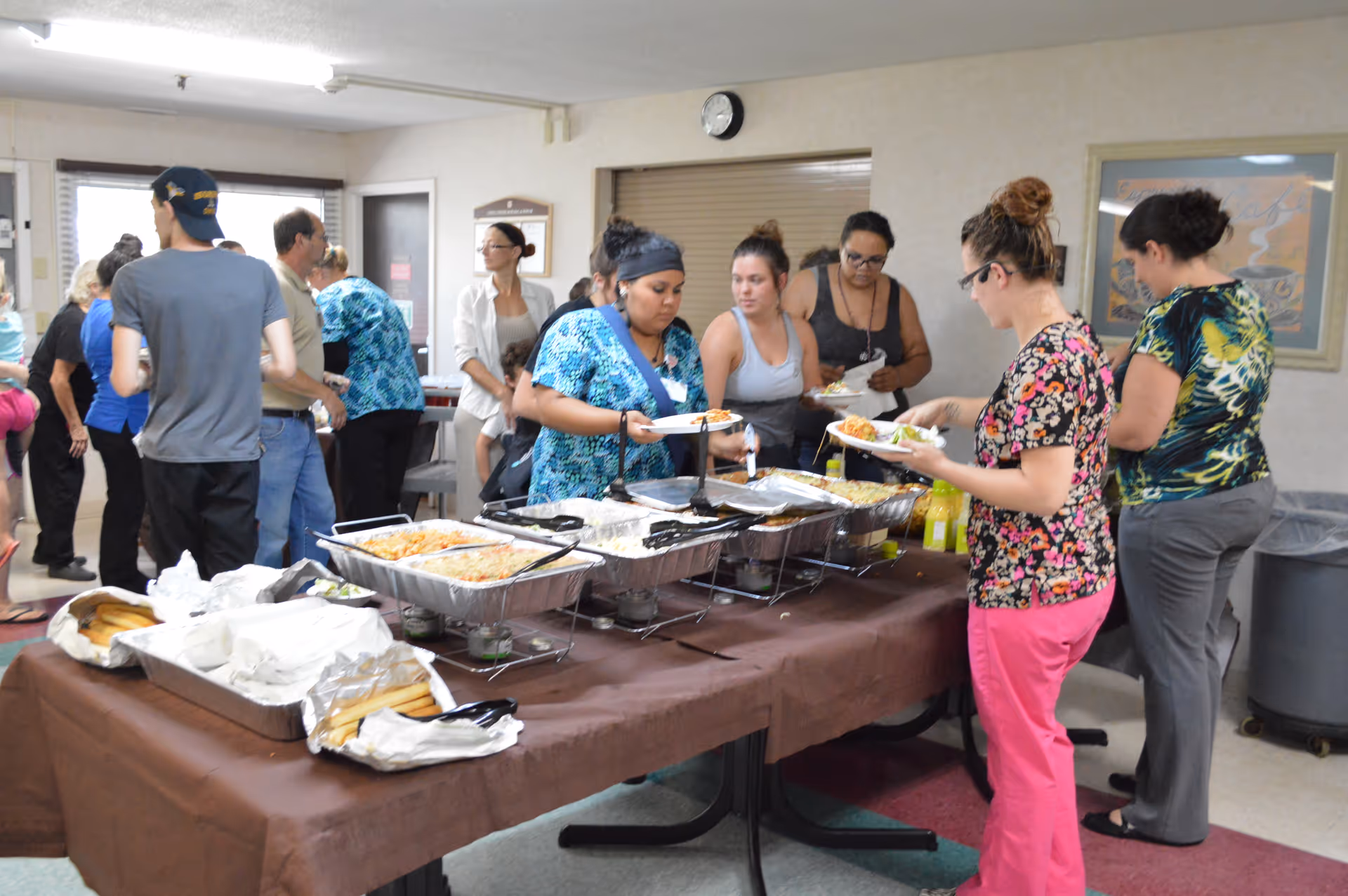 A group of people serving themselves food from a buffet table covered with a brown tablecloth in a room with beige walls. Several trays of food and plates are on the table, and people are standing in line to get food. The room has a clock on the wall and framed artwork.