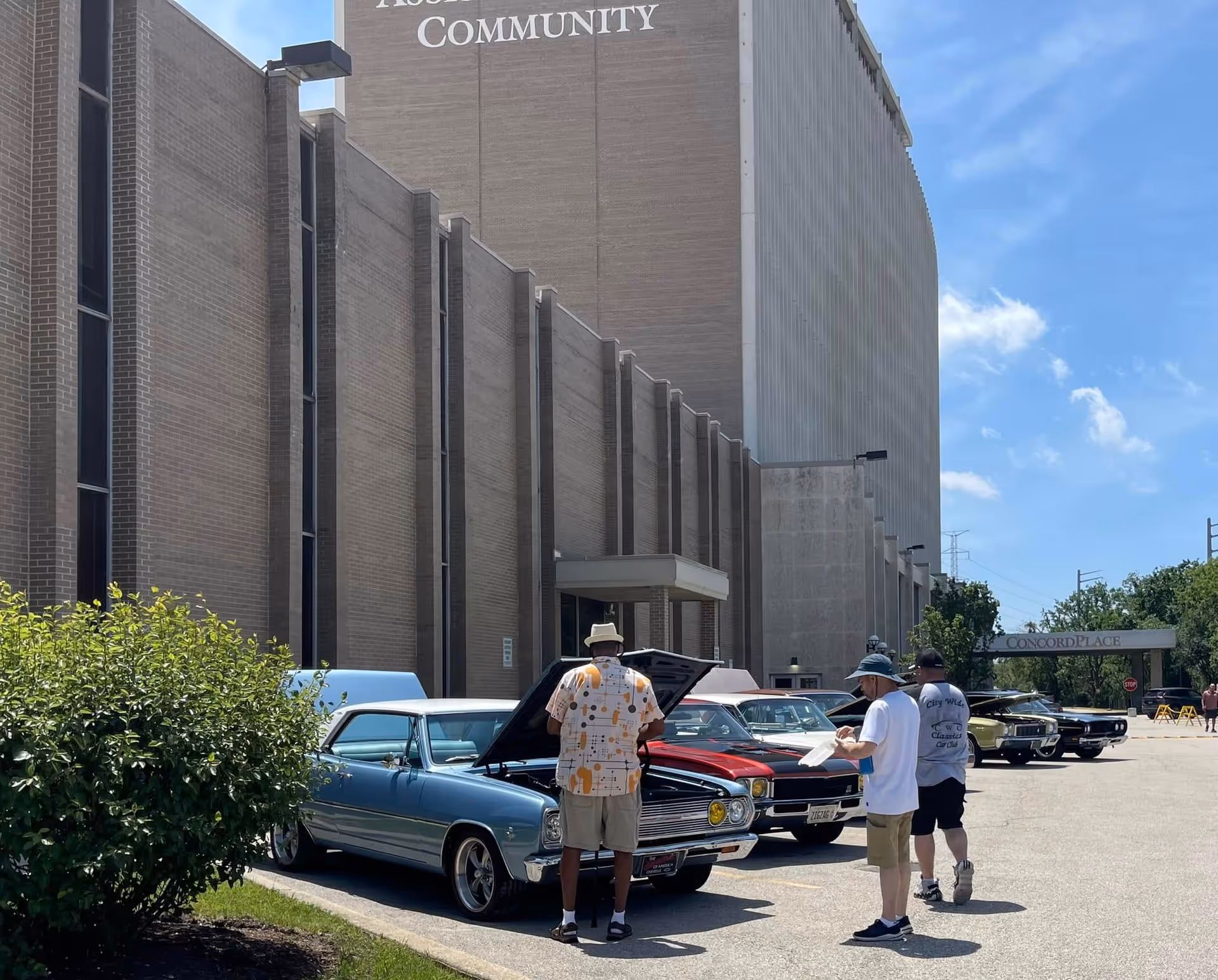 People inspecting classic cars parked outside the Concord Place Retirement Community building on a sunny day.