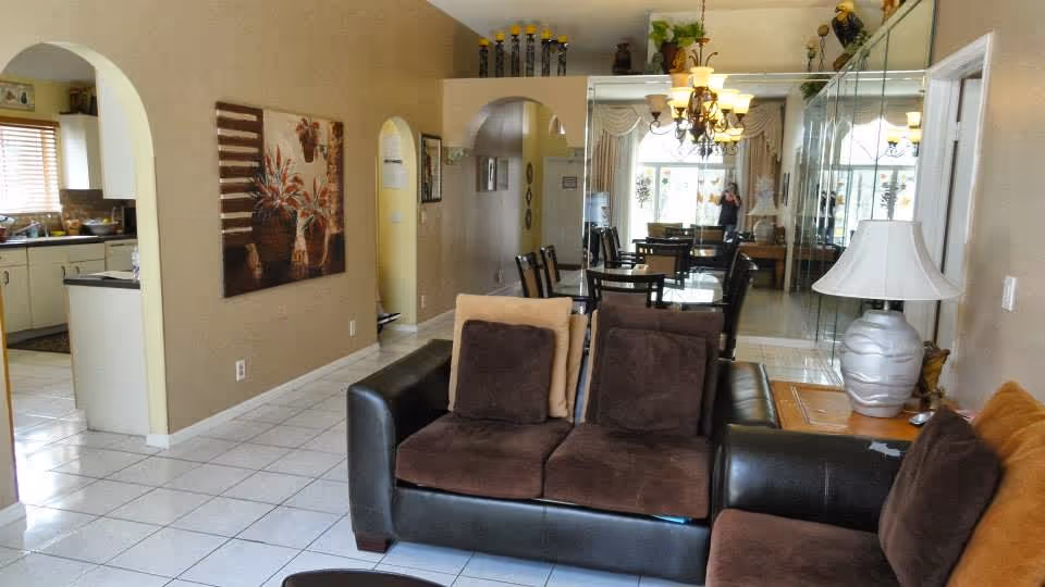 Interior view of a living room and dining area in a senior living facility. The living room has a black leather sofa with brown cushions, a side table with a white lamp, and a large painting on the wall. The dining area features a glass-top table with several chairs and a chandelier overhead. The kitchen is visible through an arched doorway on the left, and a large mirror covers the wall behind the dining table, reflecting the room and a person taking the photo.