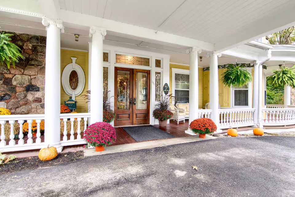 Front entrance of The Athenaeum Of Skaneateles featuring a covered porch with white columns, wooden double doors with glass panels, potted colorful flowers, pumpkins, hanging green ferns, and a stone wall on the left side.