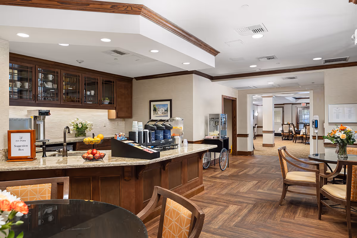 A cozy common area in a senior living facility featuring a countertop with a coffee and beverage station, a popcorn machine, and a suggestion box labeled 'Ivy Park Suggestion Box'. The room has wooden cabinetry, a vase with flowers on the counter, and several round tables with chairs. The flooring is wood-patterned, and the walls are light-colored with crown molding. In the background, there is an open doorway leading to a dining area with more tables and chairs.