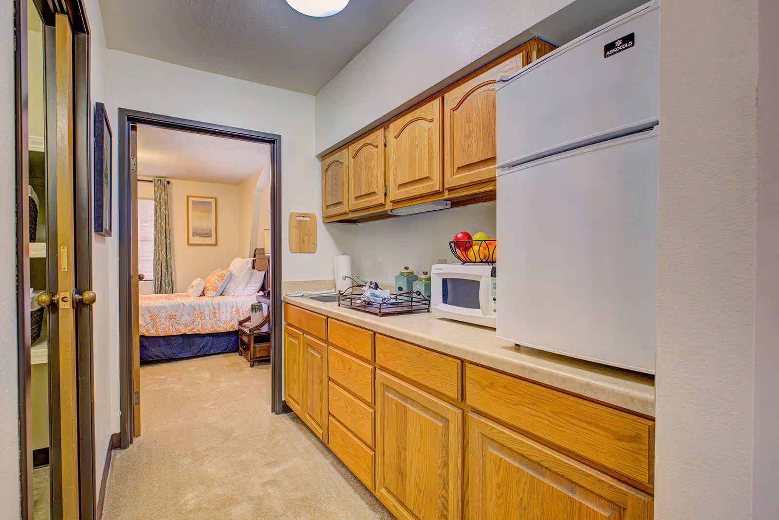 A small kitchen area with wooden cabinets, a countertop holding a microwave, a fruit basket with apples and oranges, and a white refrigerator. The kitchen opens into a bedroom with a bed covered in a floral bedspread and a framed picture on the wall.