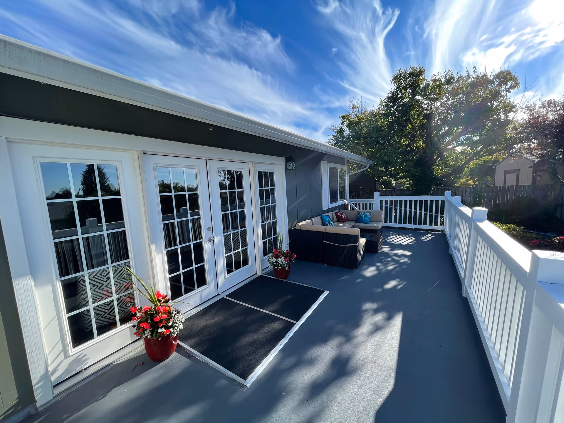 Outdoor patio area with white railing, a set of glass double doors, a potted plant with red and white flowers, and a cushioned outdoor sectional sofa with colorful pillows. Trees and a shed are visible in the background under a blue sky with wispy clouds.