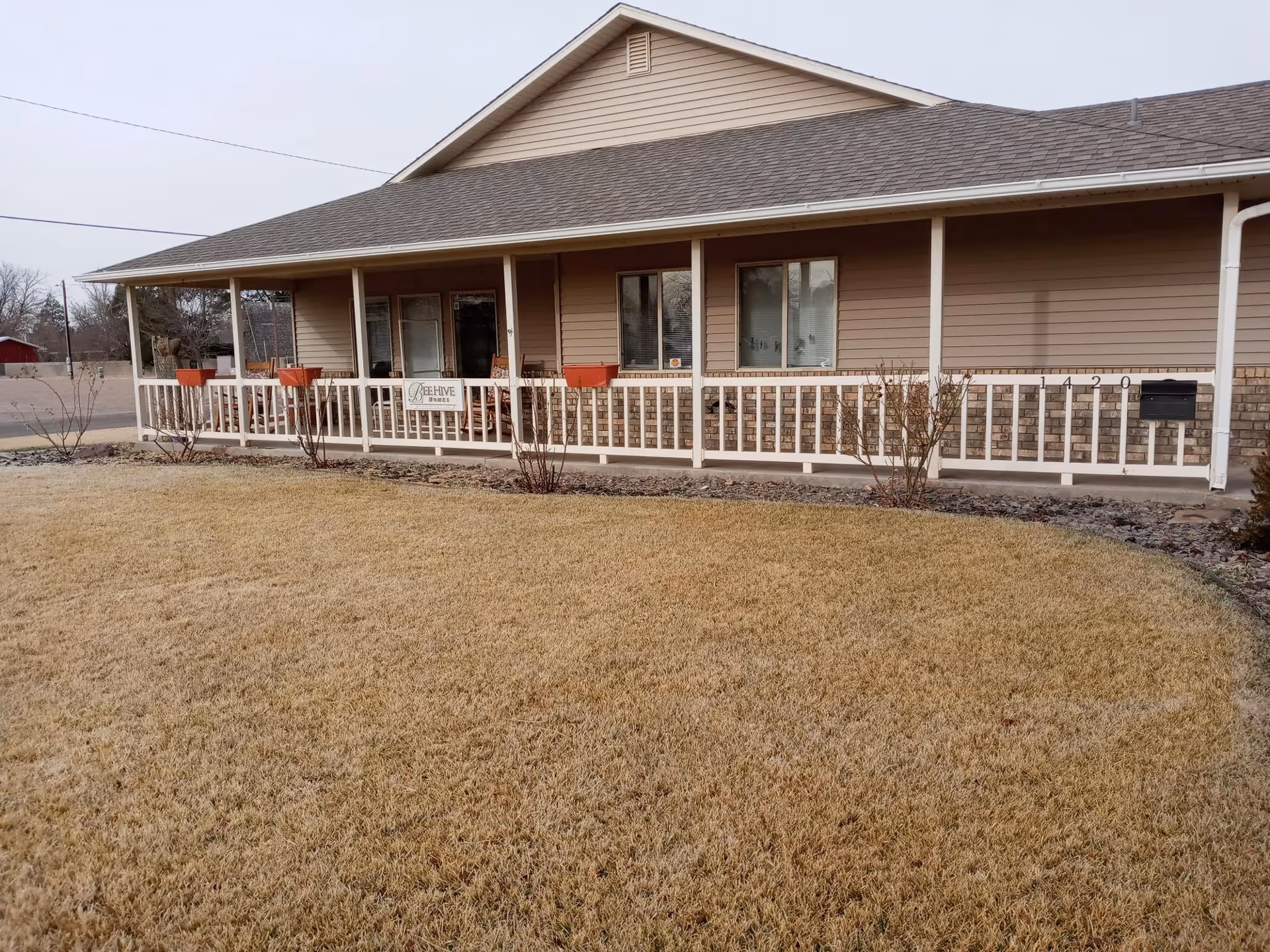 Exterior view of a single-story building with beige siding and a covered porch with white railing. There are several windows and a sign on the porch that reads 'BeeHive Homes'. The lawn in front is dry and yellowish, and there are some small bushes near the porch.