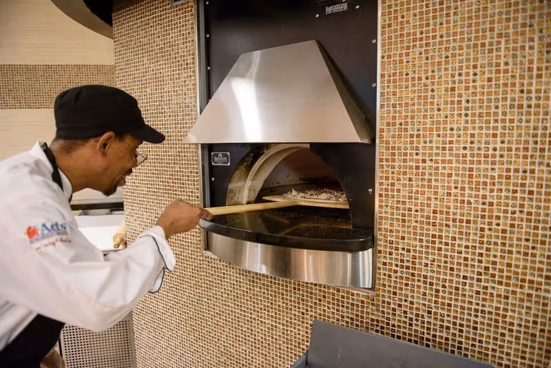 A chef wearing a white uniform and black cap is placing a pizza into a wall-mounted pizza oven with a wooden pizza peel. The oven is surrounded by small square mosaic tiles in shades of brown and beige.