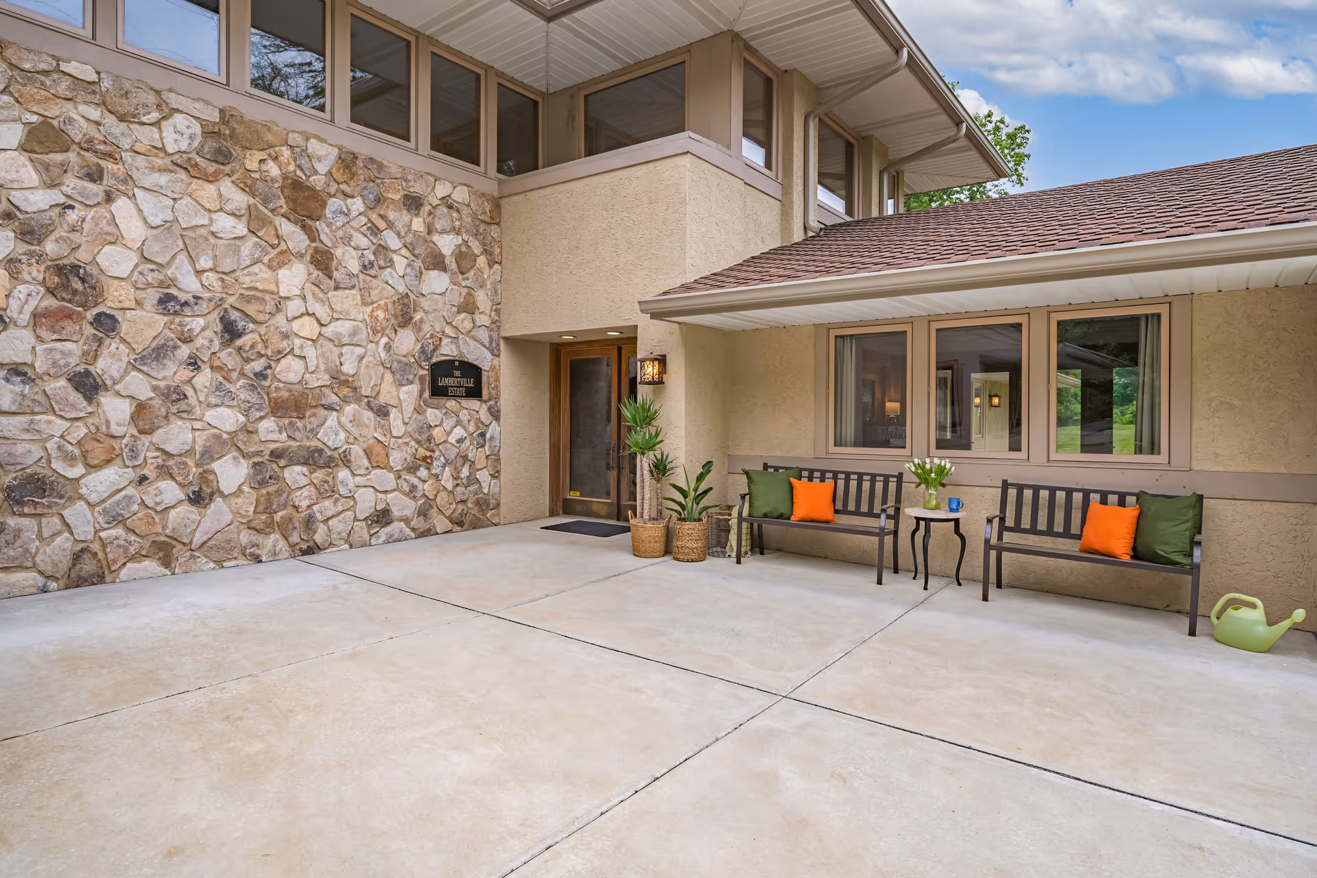 Exterior view of The Lambertville Estate entrance featuring a stone wall with a sign, a wooden door, two benches with orange and green cushions, a small table with flowers, potted plants, and a green watering can on a concrete patio under a partly cloudy sky.