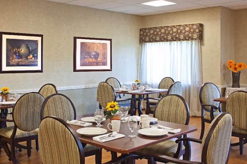 A bright dining room with multiple set tables, upholstered chairs, framed artwork, and floral centerpieces.