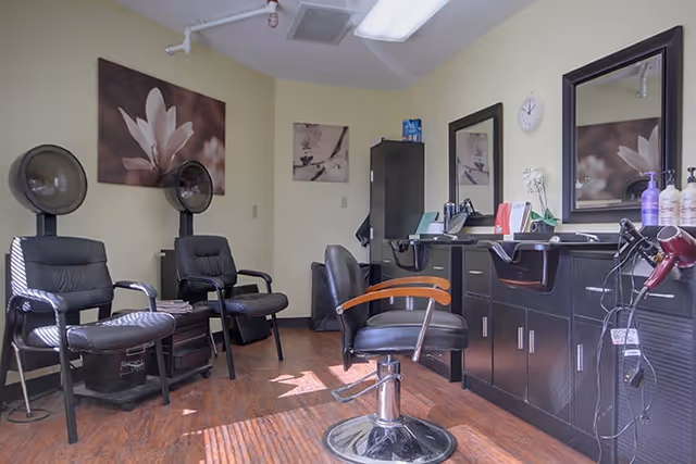 Interior of a salon area with two black hair dryer chairs on the left and a black styling chair in front of a counter with two mirrors and hair care products. The room has light yellow walls, wooden flooring, and floral artwork on the walls.