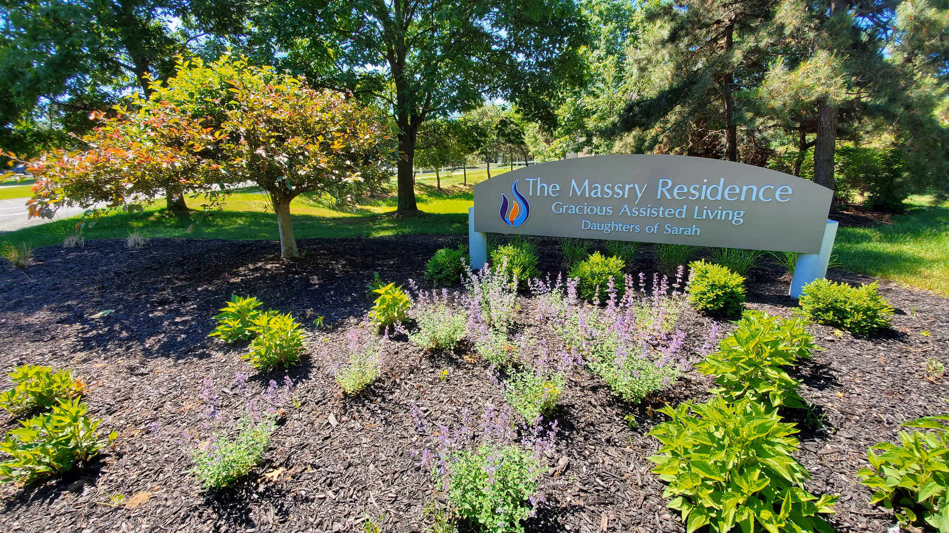 Landscaped entrance sign for The Massry Residence Gracious Assisted Living, surrounded by mulch, flowering plants and trees.