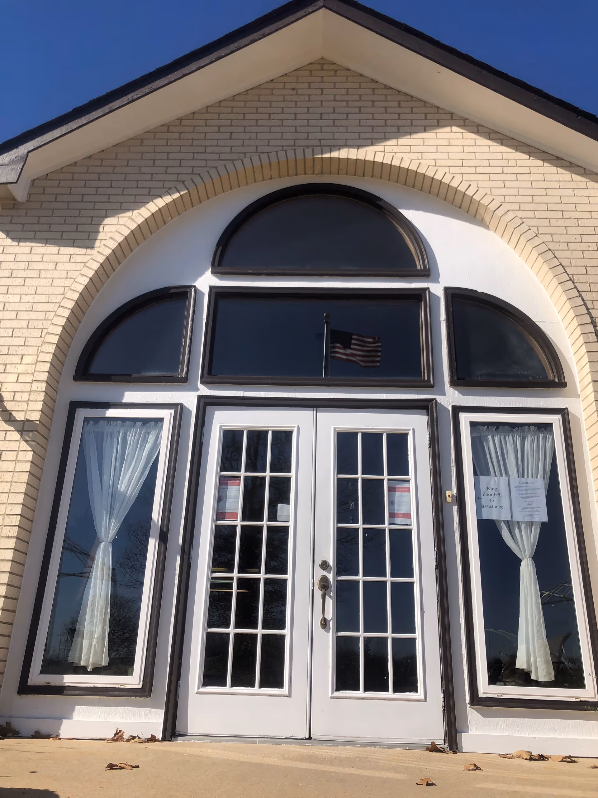Front entrance of a building with double white French doors and large arched windows above and on the sides. The building exterior is light-colored brick with a triangular roof. White curtains are visible through the side windows, and an American flag is reflected in the glass.