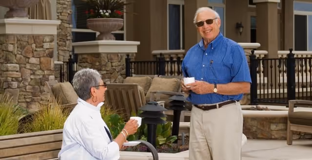An elderly man and woman enjoying a conversation outdoors at a senior living facility. The man is standing wearing sunglasses, a blue shirt, and beige pants, holding a cup and saucer. The woman is seated on a bench, wearing glasses and a white jacket, also holding a cup and saucer. The background shows stone walls, plants, and outdoor seating.