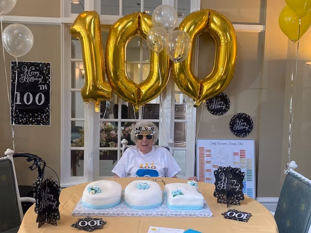 An elderly person wearing a white shirt and a birthday crown sits at a table decorated for a 100th birthday celebration. Large gold balloons form the number 100 above the person. The table has a cake shaped as the number 100 with white and blue icing. There are black and white 'Happy Birthday' signs and balloons around the room.