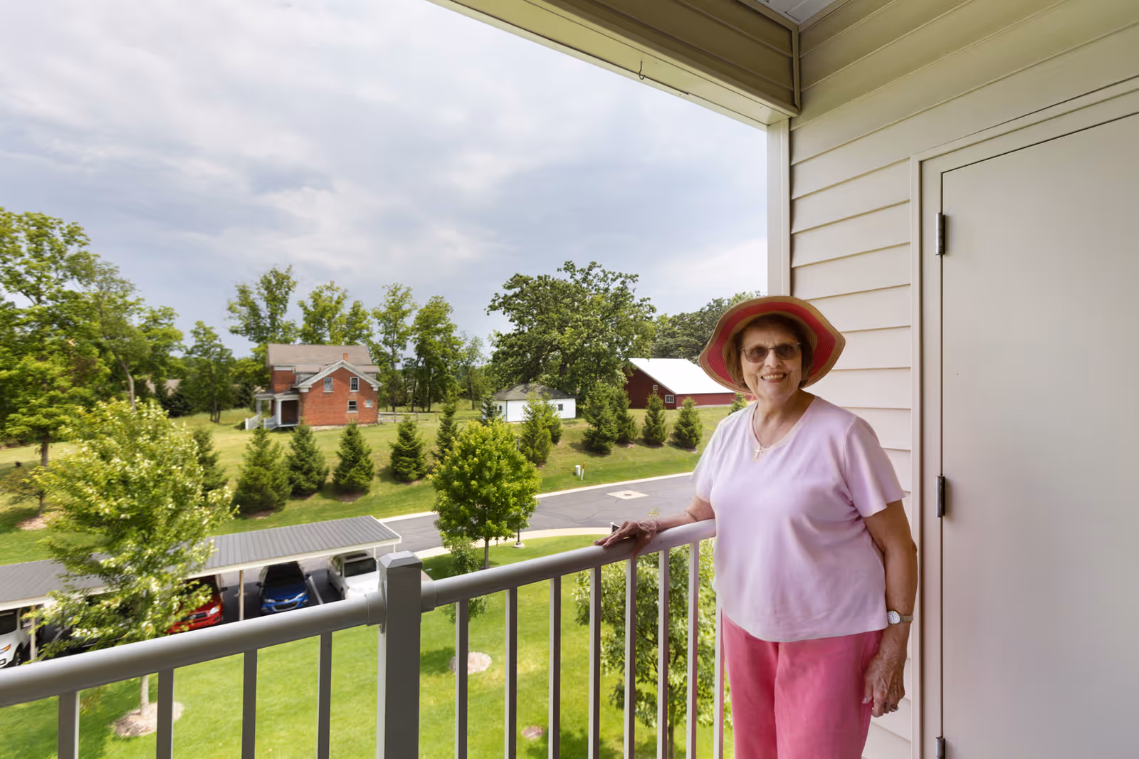 A woman in a pink outfit and sunhat stands on a balcony overlooking green lawns, parked cars, and houses.