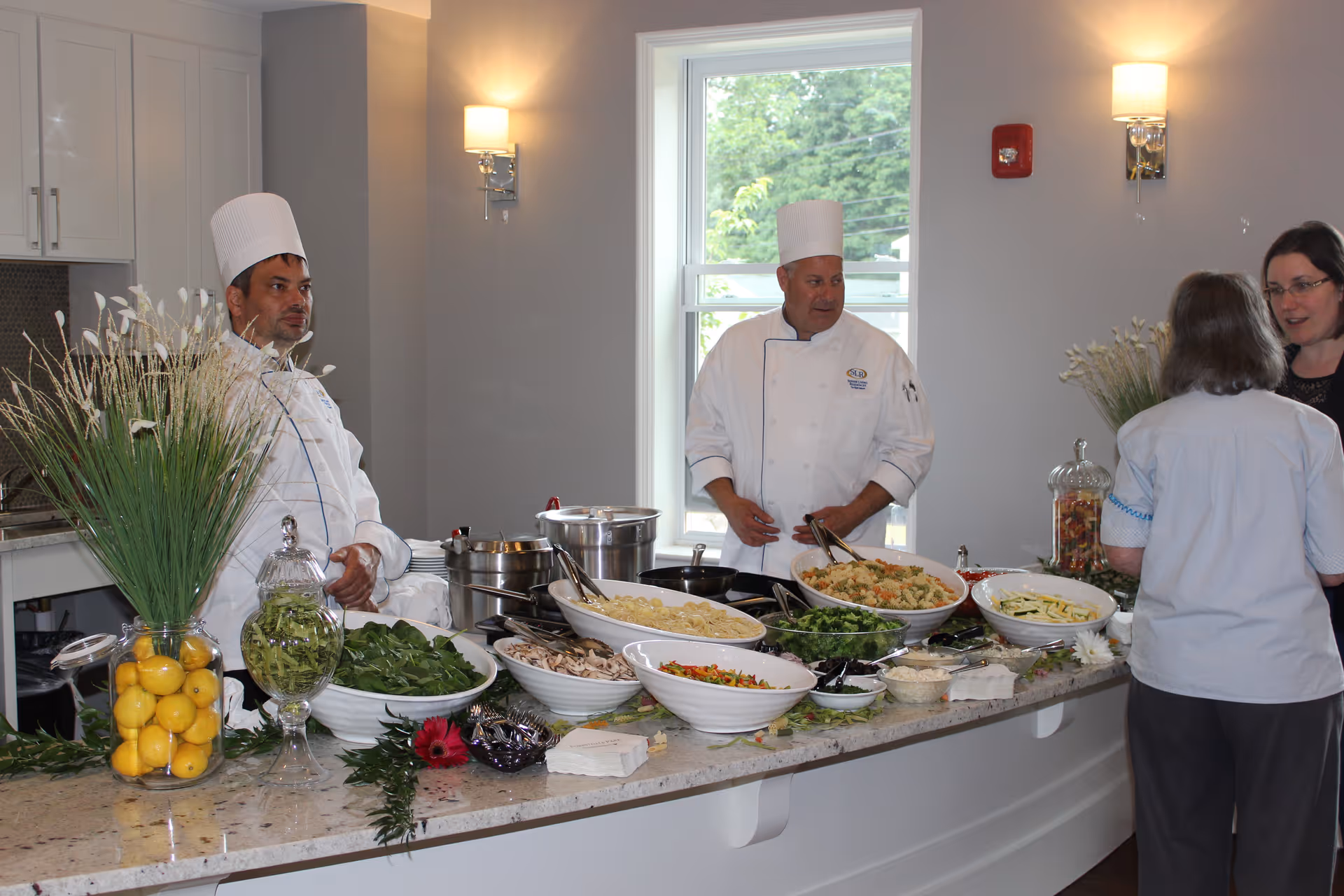 Two chefs in white uniforms and hats stand behind a counter filled with bowls of fresh vegetables, pasta, and salad ingredients. Two women stand on the opposite side of the counter, engaged in conversation. The setting appears to be a bright kitchen or dining area with white cabinets, a window, and wall-mounted lights.