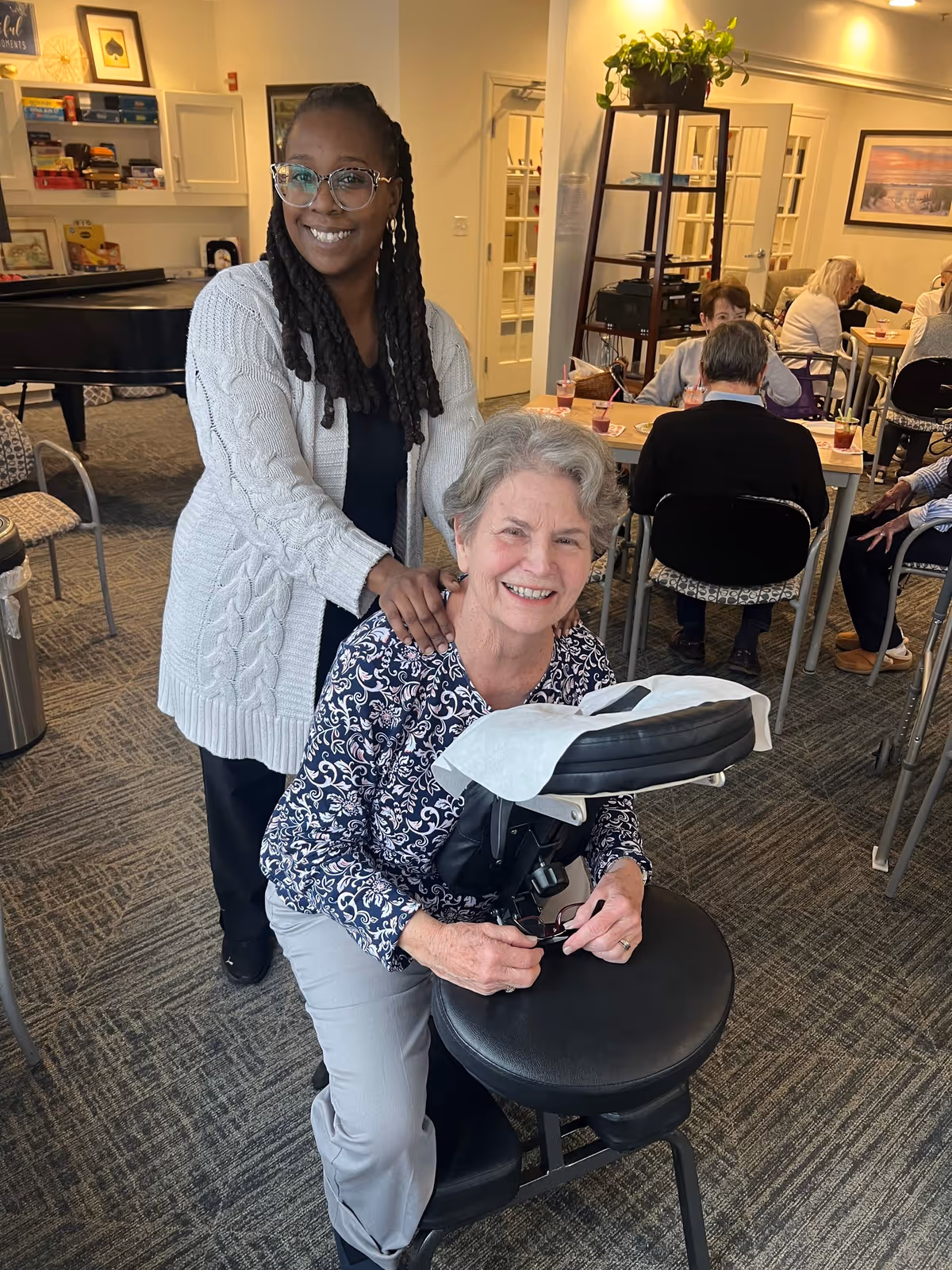 A smiling elderly woman is seated on a massage chair receiving a shoulder massage from a younger woman wearing glasses and a white cardigan. In the background, several elderly people are seated at tables in a well-lit room with carpeted floors and framed artwork on the walls.