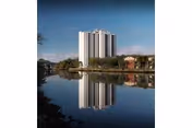 Tall white apartment building reflected in a calm body of water with trees and smaller buildings along the shore under a blue sky.