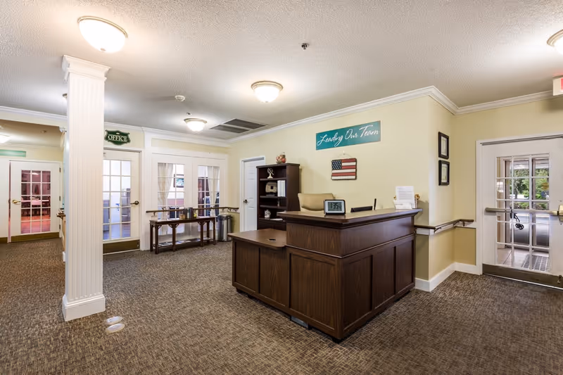 Reception area inside Sodalis Tallahassee Assisted Living facility with a wooden front desk, a digital clock, and a sign on the wall that reads 'Leading Our Team'. There are multiple doors with glass panels, a small table with items on it, and a carpeted floor. The area is well-lit with ceiling lights.